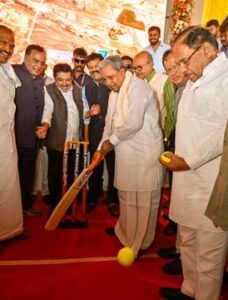 Karnataka Chief Minister Siddaramaiah plays a symbolic cricket shot with a bat during a public function, interacting with attendees at the event.